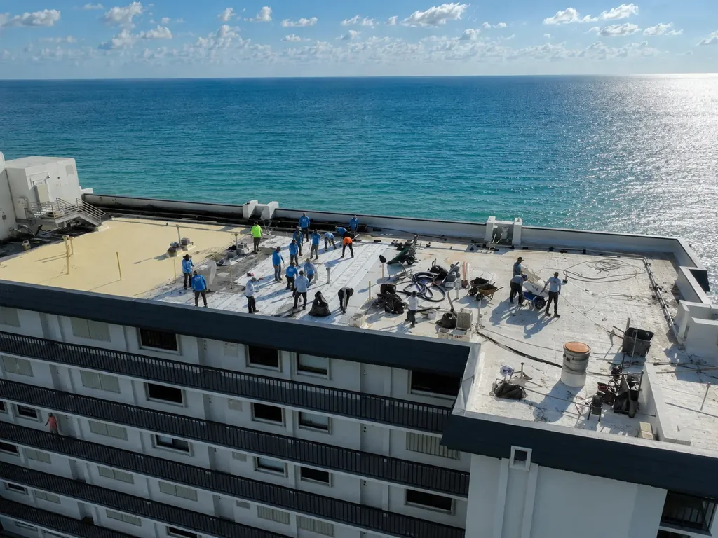 Aerial view of an SCI Roofing crew spread across the flat roof of an oceanfront building, membrane rolled out over part of the deck and the Atlantic filling the frame behind them